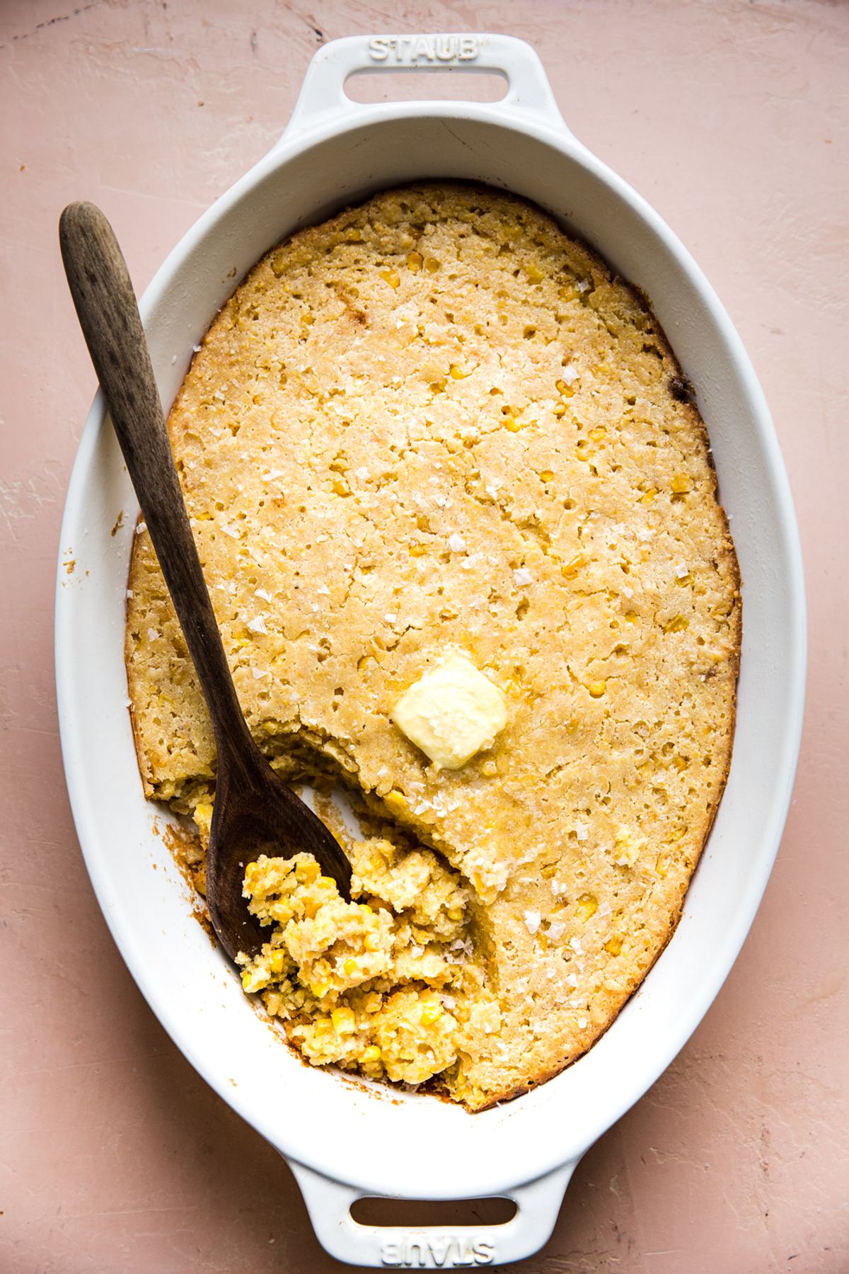 fresh baked spoon bread in a casserole dish topped with a pat of butter and being scooped out with a wooden spoon